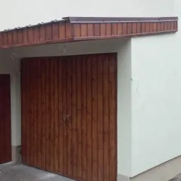 Biała elewacja budynku z drewnianą bramą garażową, widoczny fragment dachu i popękana posadzka przed budynkiem