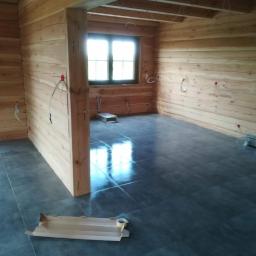 Usługi remontowo budowlane - Surowe wnętrze domu z bali z ciemną, błyszczącą podłogą z płytek, widoczne kable elektryczne wystające ze ścian i kartonowe elementy na podłodze.