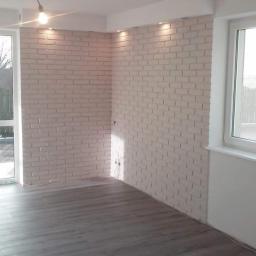 Usługi remontowo budowlane - Wykończone wnętrze z imitacją cegły na ścianach w odcieniach bieli, szara podłoga panelowa, sufit z oświetleniem punktowym i widocznym przewodem lampy wiszącej, okno z widokiem na zewnątrz.