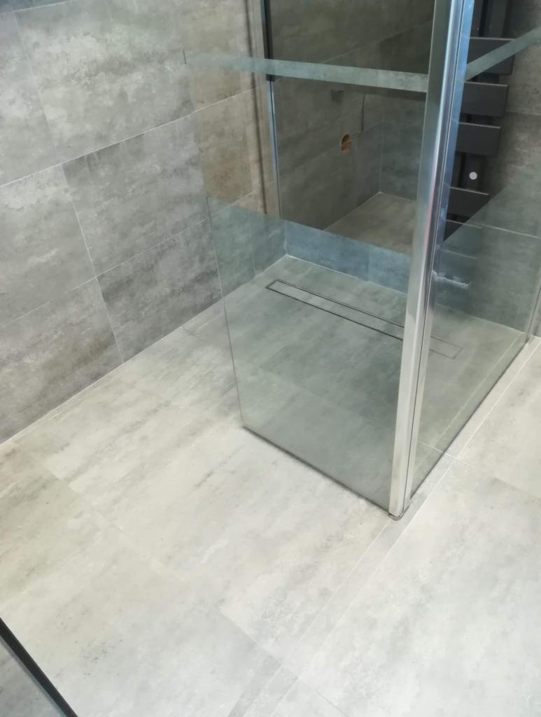 Nowoczesna kabina prysznicowa z odpływem liniowym w posadzce, wykończona szarymi płytkami imitującymi kamień, z widocznym grzejnikiem drabinkowym w tle.