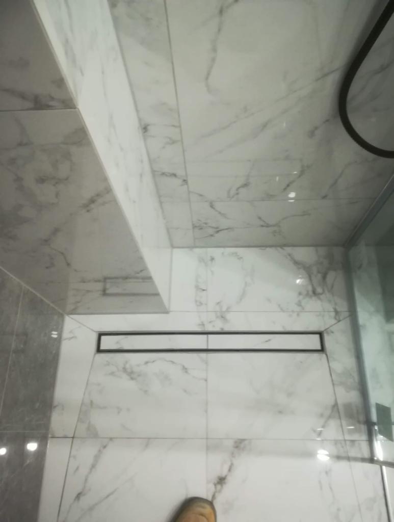 Nowoczesna łazienka z marmurowymi płytkami, czarnym odpływem liniowym pod prysznicem i fragmentem kabiny prysznicowej.