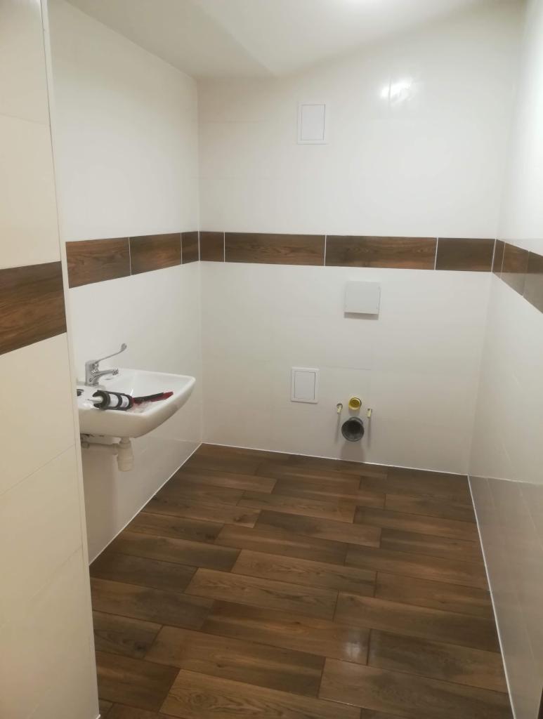 Wnętrze łazienki w trakcie prac wykończeniowych, widoczna biała umywalka z baterią, brązowe płytki imitujące drewno na podłodze i pas płytek na ścianie, instalacja kanalizacyjna wystająca ze ściany.