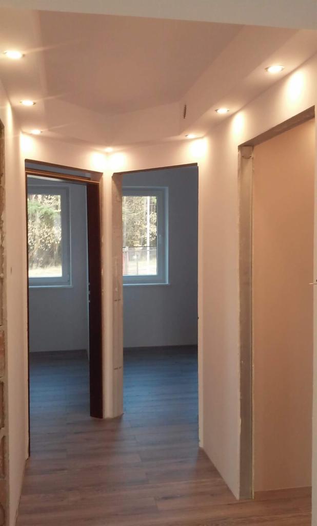 Wnętrze mieszkania w trakcie prac wykończeniowych, widoczne osadzone ościeżnice drzwiowe, podłoga wyłożona panelami imitującymi drewno i zamontowane oświetlenie sufitowe punktowe.