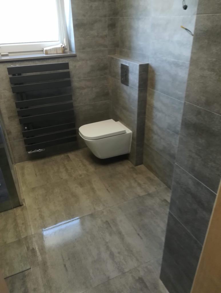 Nowoczesna łazienka w odcieniach szarości z podwieszaną toaletą, grzejnikiem drabinkowym i dużymi płytkami imitującymi kamień.