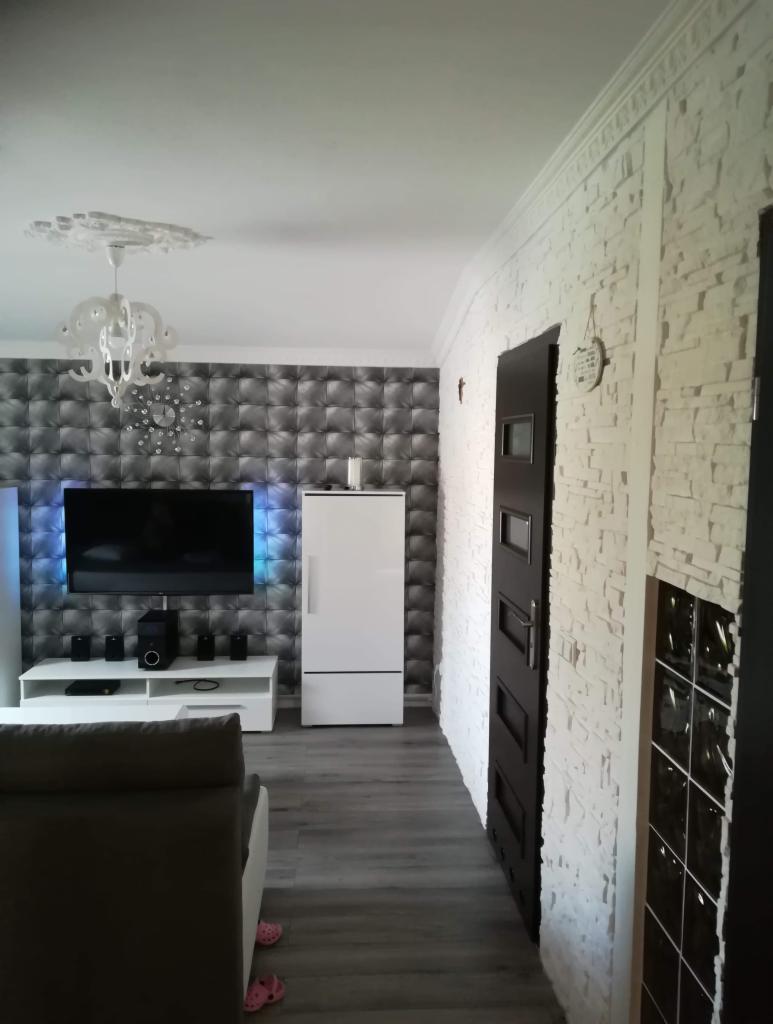 Salon po metamorfozie: ściana z dekoracyjnej cegły w odcieniach bieli, tapeta 3D w geometryczny wzór, nowoczesne oświetlenie LED za telewizorem, białe meble i szara podłoga imitująca drewno.