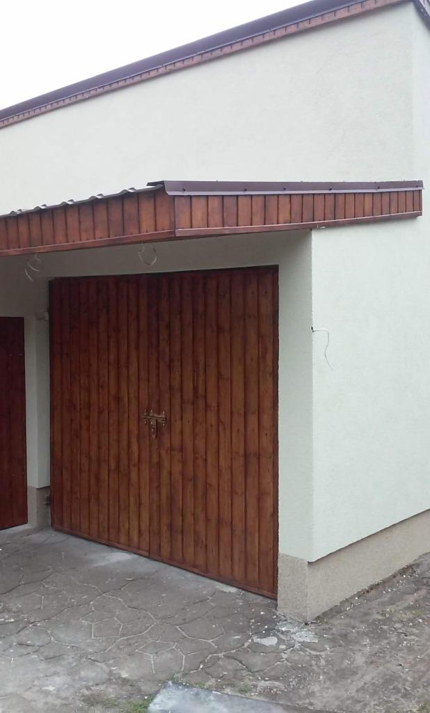 Biała elewacja budynku z drewnianą bramą garażową, widoczny fragment dachu i popękana posadzka przed budynkiem