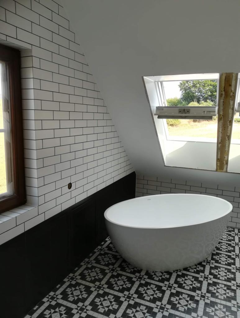 Nowoczesna łazienka na poddaszu z wolnostojącą białą wanną, biało-czarną podłogą w geometryczne wzory oraz ścianami wyłożonymi białymi płytkami imitującymi cegłę z czarną fugą, okno dachowe...