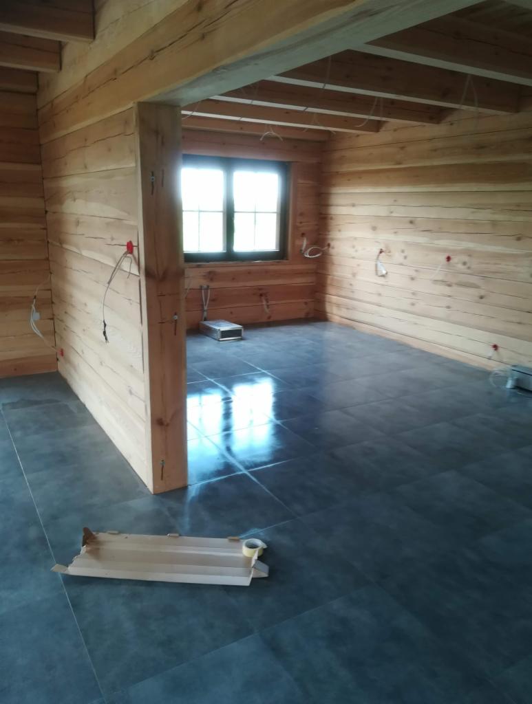 Surowe wnętrze domu z bali z ciemną, błyszczącą podłogą z płytek, widoczne kable elektryczne wystające ze ścian i kartonowe elementy na podłodze.