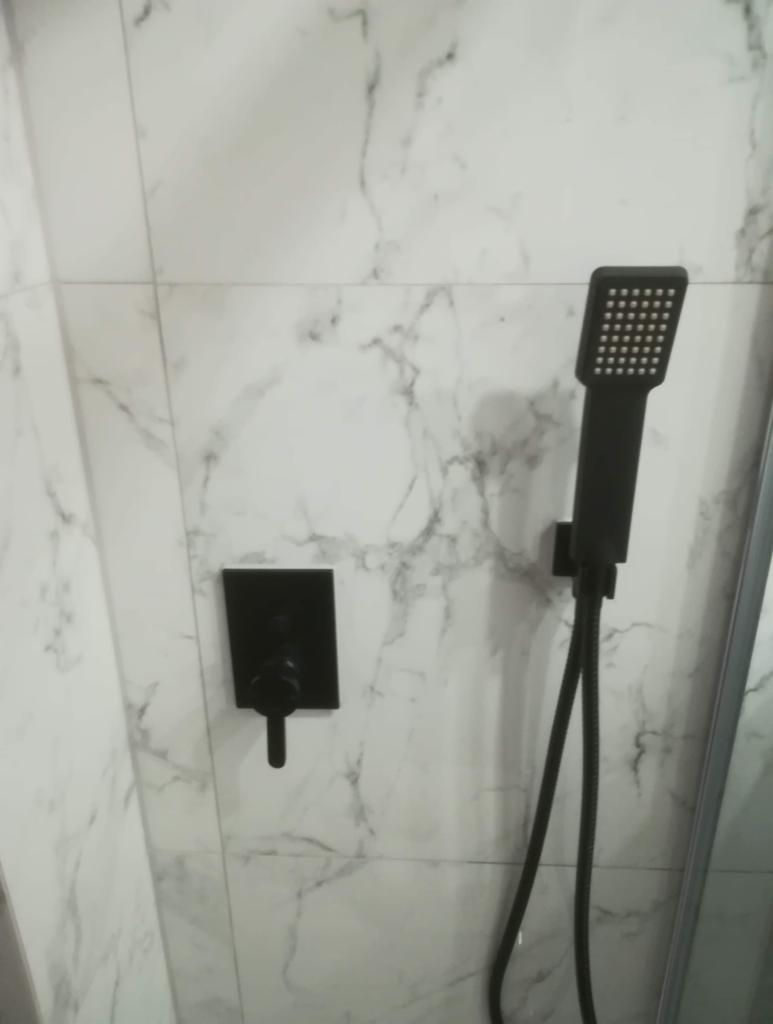 Czarna bateria natryskowa z deszczownicą i termostatem na ścianie wyłożonej białymi płytkami imitującymi marmur w kabinie prysznicowej.