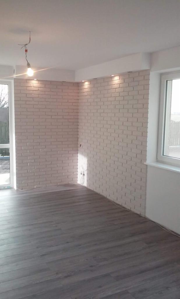 Wykończone wnętrze z imitacją cegły na ścianach w odcieniach bieli, szara podłoga panelowa, sufit z oświetleniem punktowym i widocznym przewodem lampy wiszącej, okno z widokiem na zewnątrz.