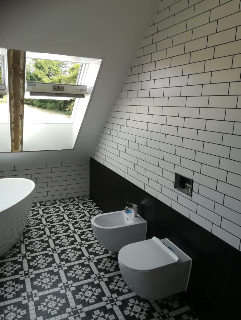Wykończona łazienka na poddaszu z biało-czarną mozaiką na podłodze, białą wanną, bidetem i wiszącą toaletą, ściany wyłożone białymi płytkami imitującymi cegłę z czarnymi fugami, okno dachowe...
