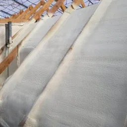 Skośny dach od wewnątrz, pokryty grubą warstwą białej pianki izolacyjnej, widoczne drewniane krokwie i konstrukcja dachu