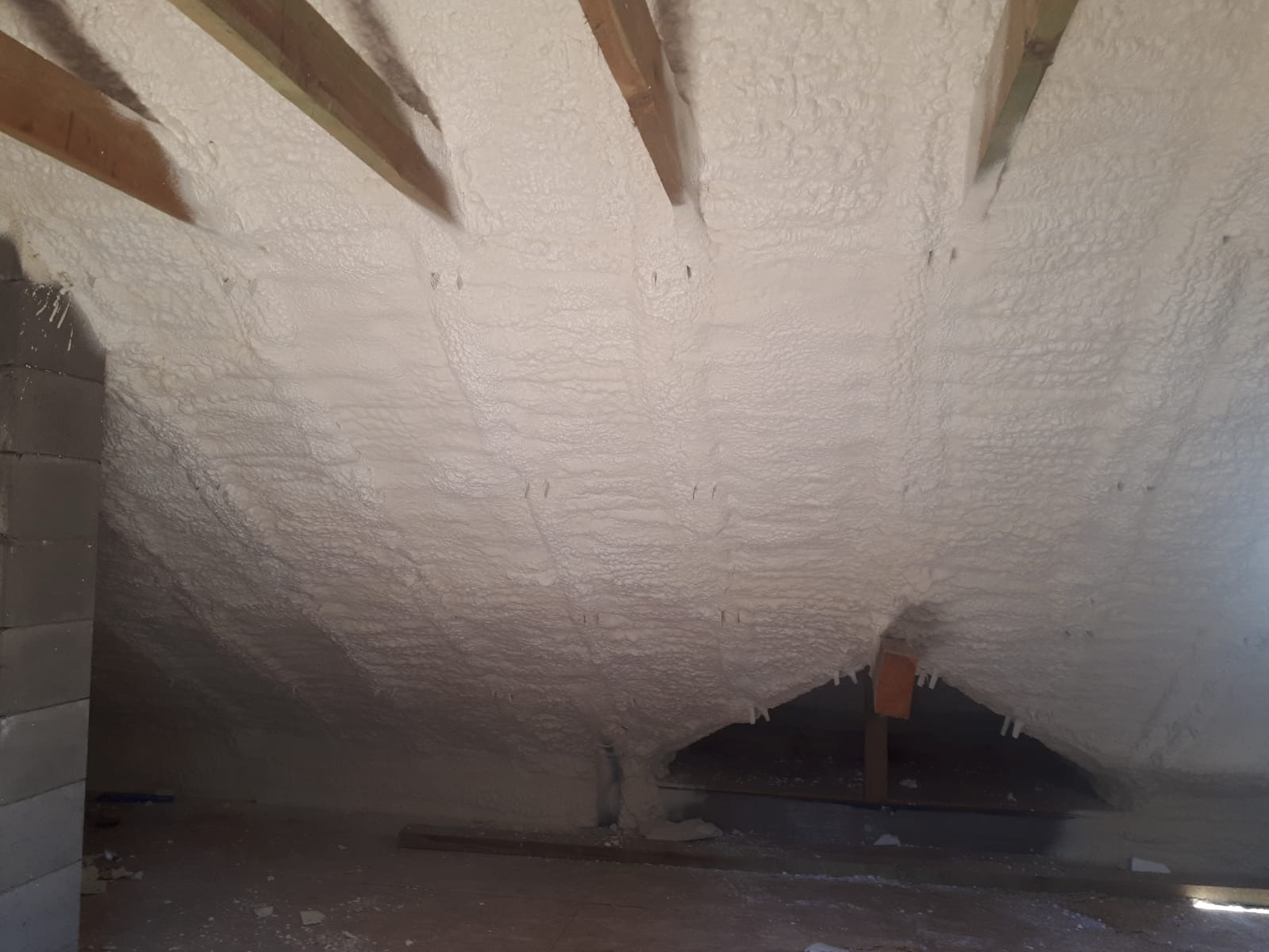 Poddasze z widocznymi elementami konstrukcji drewnianej, pokryte grubą warstwą białej izolacji natryskowej.