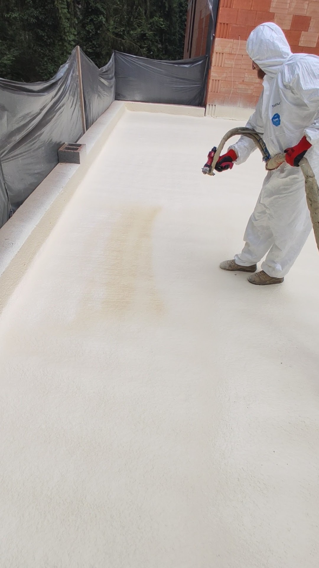 Pracownik w kombinezonie ochronnym aplikuje izolację natryskową na płaski dach w Konstancin-Jeziorna, widoczny fragment muru i zabezpieczona folią przestrzeń.
