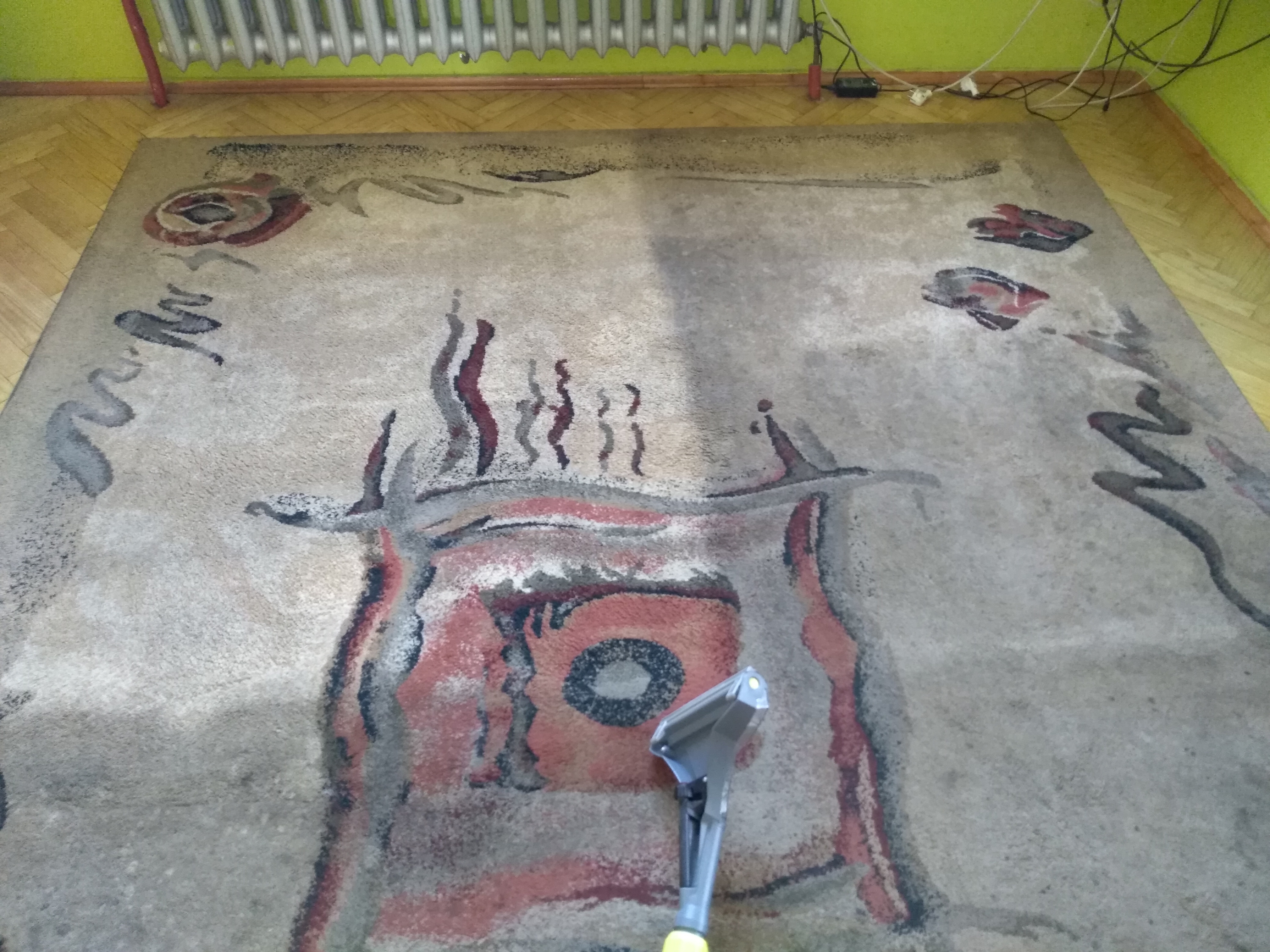 Dysza odkurzacza piorącego na wzorzystym dywanie, widoczny fragment wyczyszczonej powierzchni, parkiet i grzejnik w tle.
