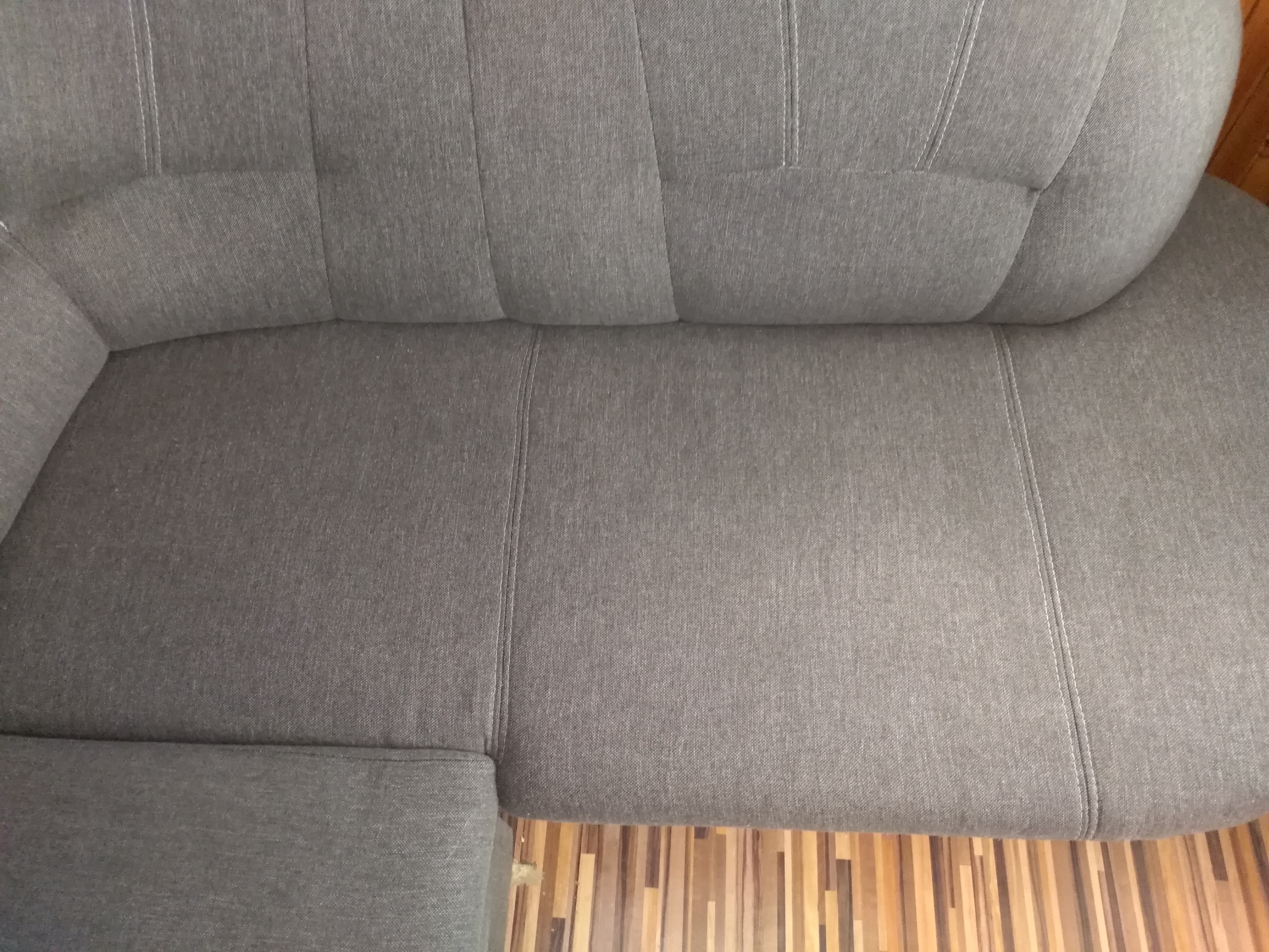Fragment szarej sofy z widocznymi szwami i fakturą materiału, ujęcie z góry, na tle drewnianej podłogi.