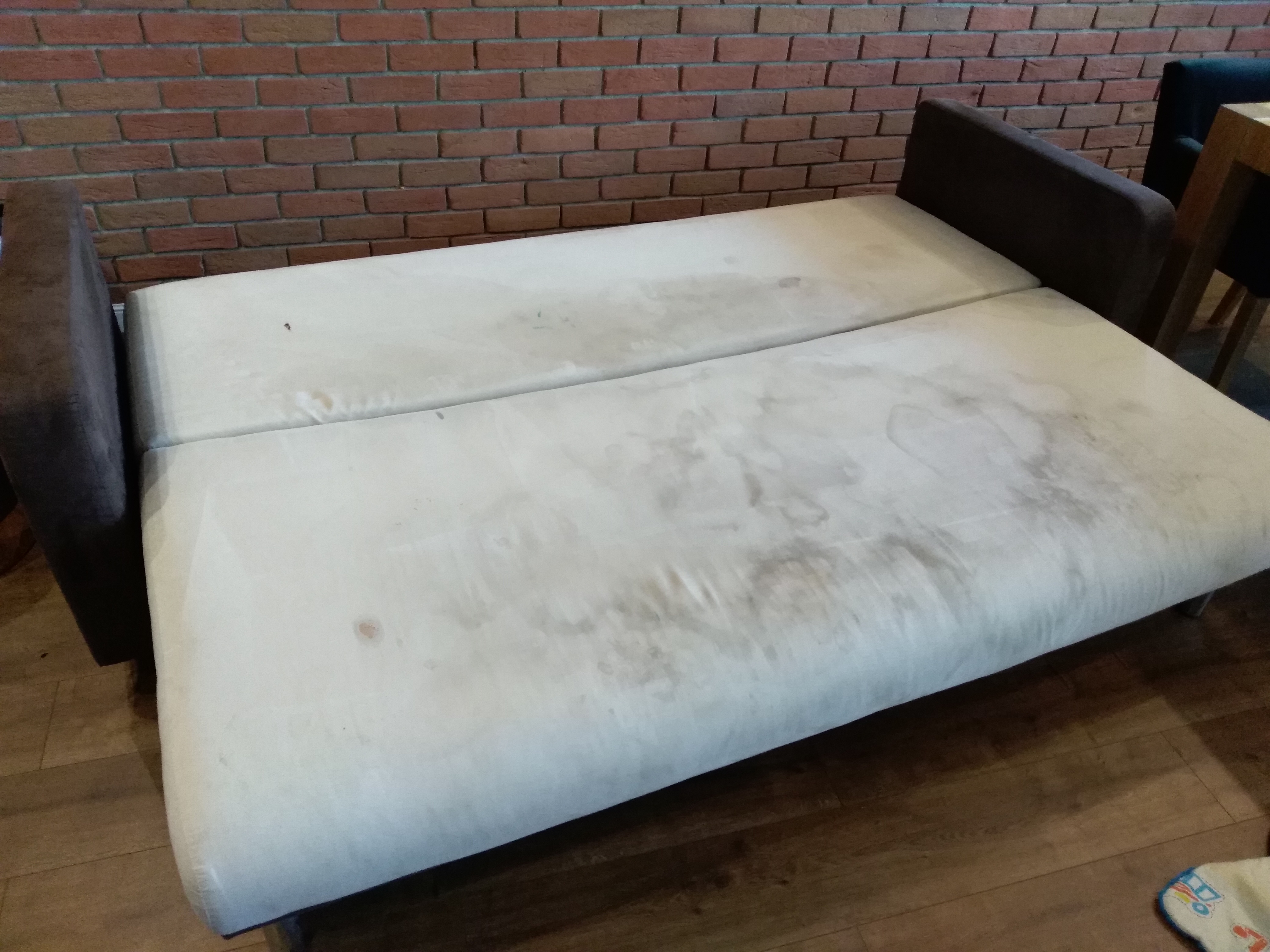 Rozłożona, jasna kanapa z widocznymi plamami na tapicerce, ustawiona na drewnianej podłodze przed ścianą z czerwonej cegły.