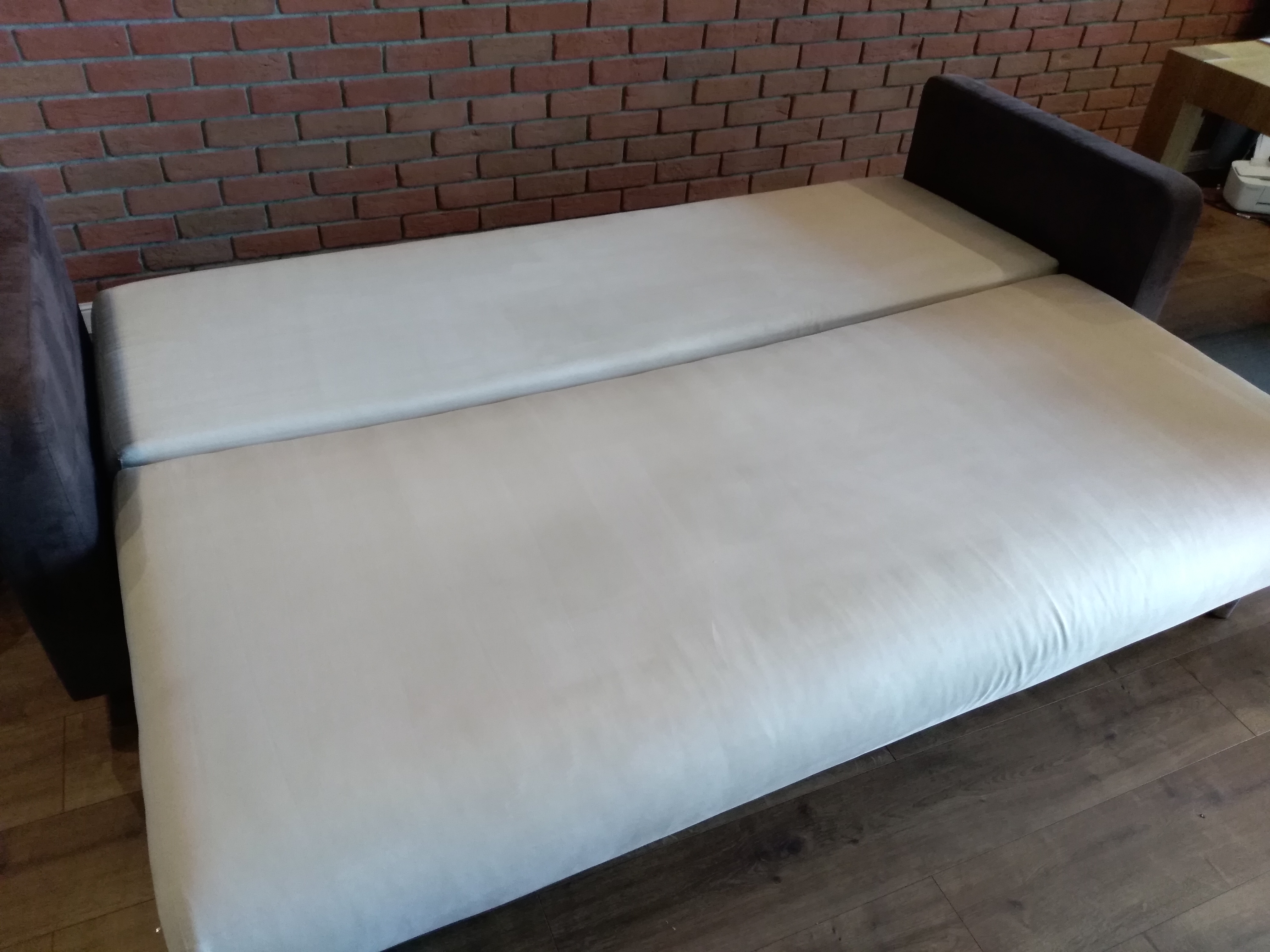 Rozłożona sofa z jasną tapicerką na tle ceglanej ściany i drewnianej podłogi.