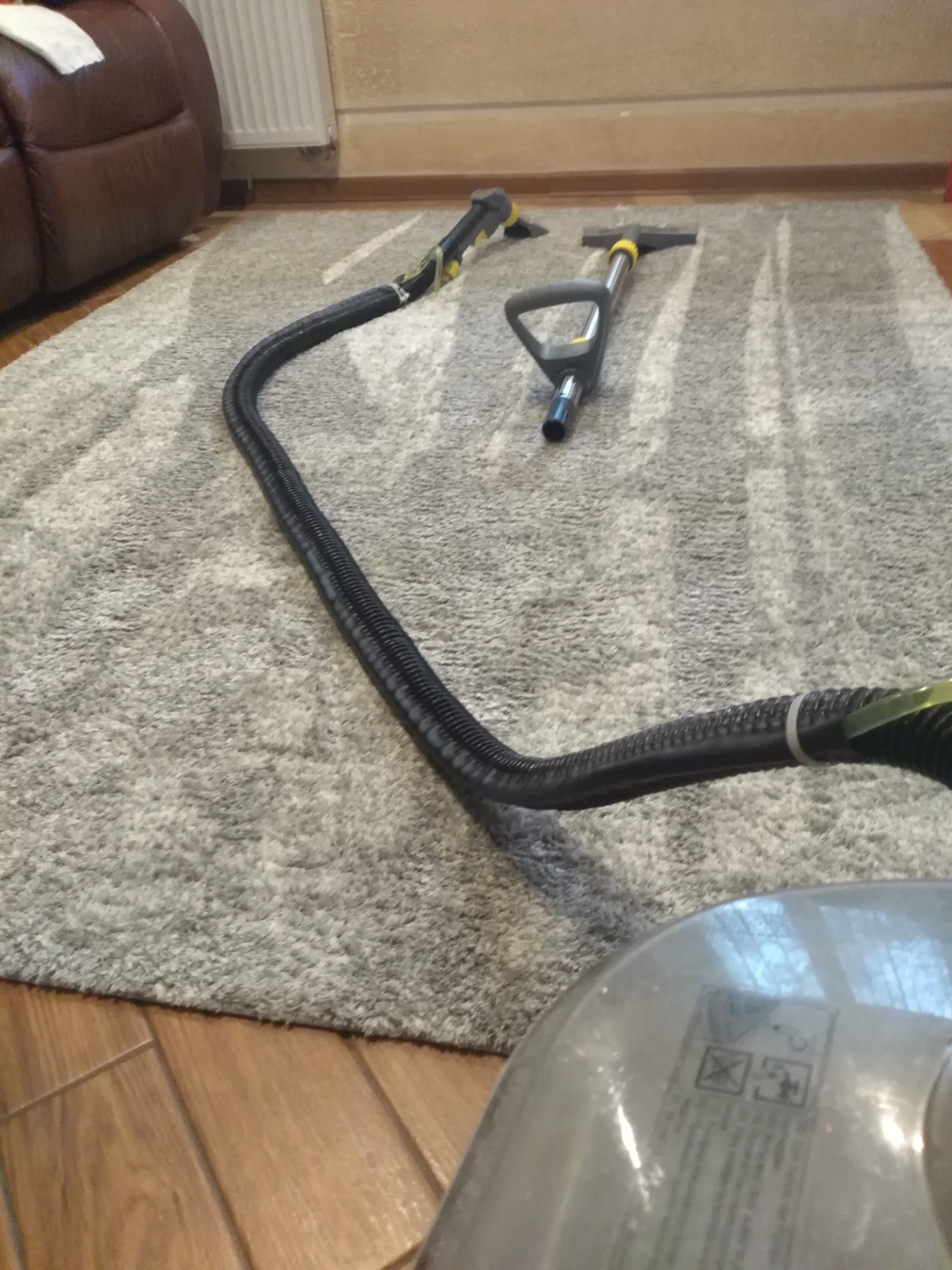 Dysze i wąż odkurzacza piorącego leżące na szarym dywanie z widocznymi śladami czyszczenia, obok widoczny fragment urządzenia.