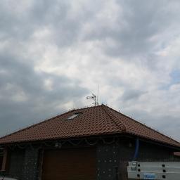 Voltage Adam Kwaśniewski - Dom jednorodzinny z brązową dachówką, anteną telewizyjną na dachu i widocznym systemem odgromowym, drabina Krause w tle, pochmurne niebo.