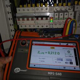 Voltage Adam Kwaśniewski - Miernik Sonel MPI-540 z wyświetlonym wynikiem pomiaru impedancji pętli zwarcia ZL-PE = 0,212 Ω, na tle rozdzielnicy elektrycznej z wyłącznikami nadprądowymi i różnicowoprądowymi.