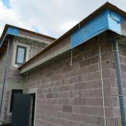 Voltage Adam Kwaśniewski - Surowy budynek z szarej cegły z widoczną instalacją elektryczną na elewacji i niebieskim zabezpieczeniem dachu, przewody poprowadzone po zewnętrznej ścianie.