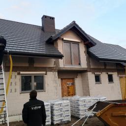 Voltage Adam Kwaśniewski - Montaż instalacji odgromowej na nowym domu z szarej dachówki, widoczny pracownik na drabinie i stosy materiałów budowlanych przed budynkiem.