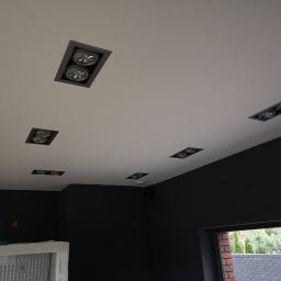 Voltage Adam Kwaśniewski - Biały sufit z zainstalowanymi, kwadratowymi oprawami oświetleniowymi z podwójnymi żarówkami LED, ciemne ściany w tle i okno z widokiem na zewnątrz.