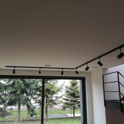 Voltage Adam Kwaśniewski - Nowoczesne oświetlenie szynowe z czarnymi reflektorami zamontowane na suficie w jasnym wnętrzu z dużym oknem i widokiem na drzewa oraz schodami z metalową balustradą.