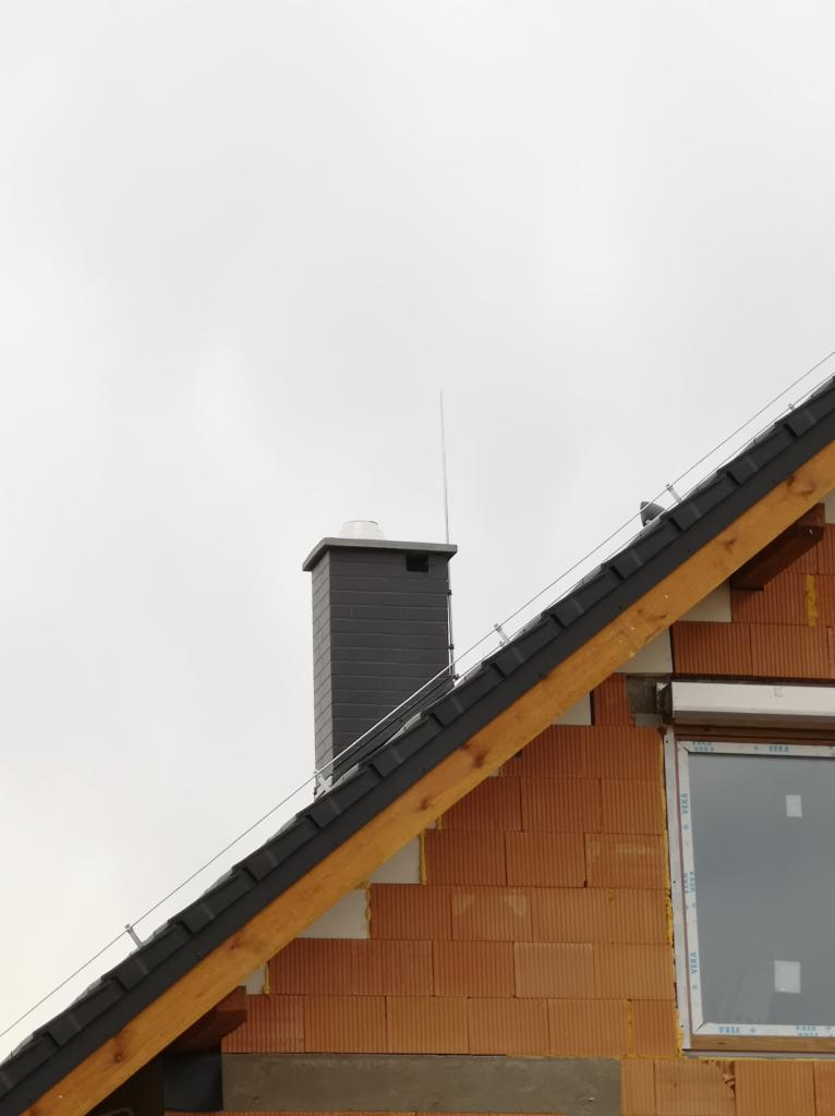 Szary komin i system ochrony odgromowej na dachu w trakcie budowy, widoczne cegły ceramiczne i drewniana konstrukcja więźby dachowej.