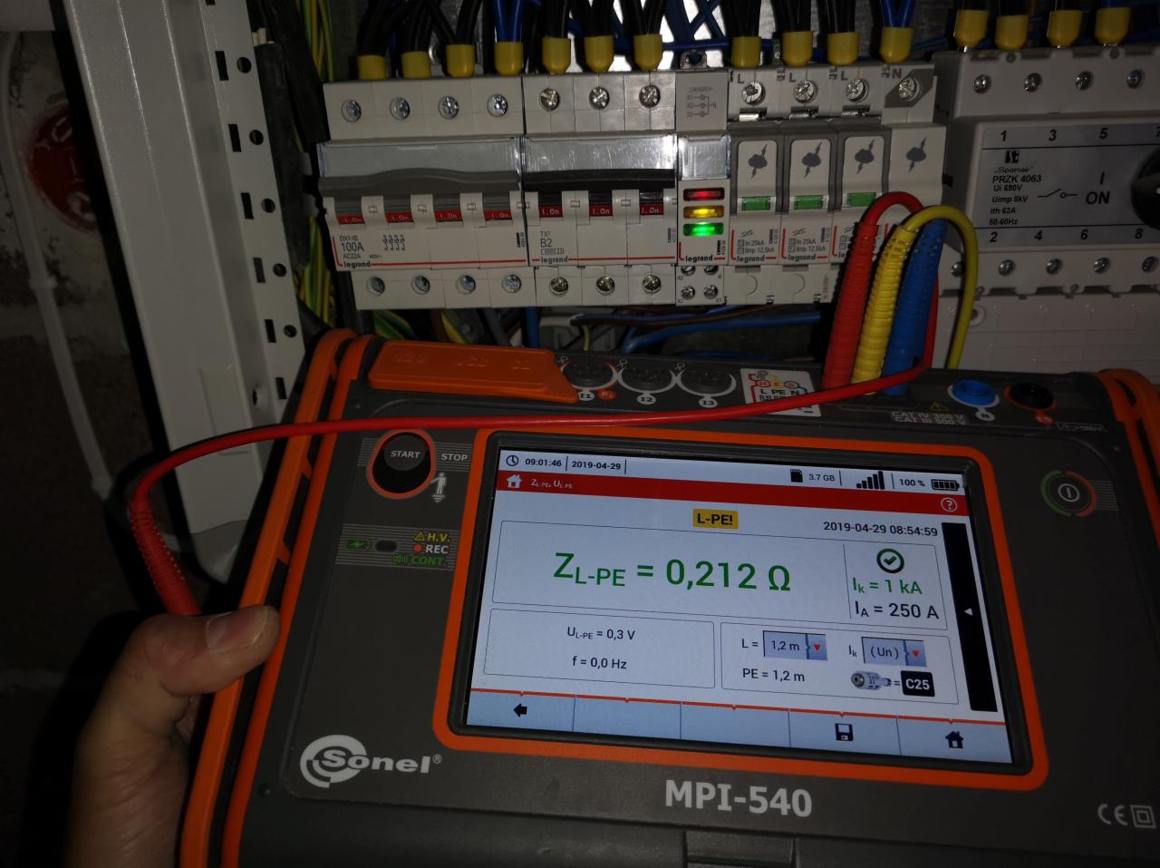 Miernik Sonel MPI-540 z wyświetlonym wynikiem pomiaru impedancji pętli zwarcia ZL-PE = 0,212 Ω, na tle rozdzielnicy elektrycznej z wyłącznikami nadprądowymi i różnicowoprądowymi.
