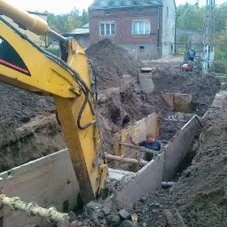 Żółta koparka CAT umieszcza betonowy element w wykopie zabezpieczonym szalunkiem, w tle budynki i hałda ziemi.