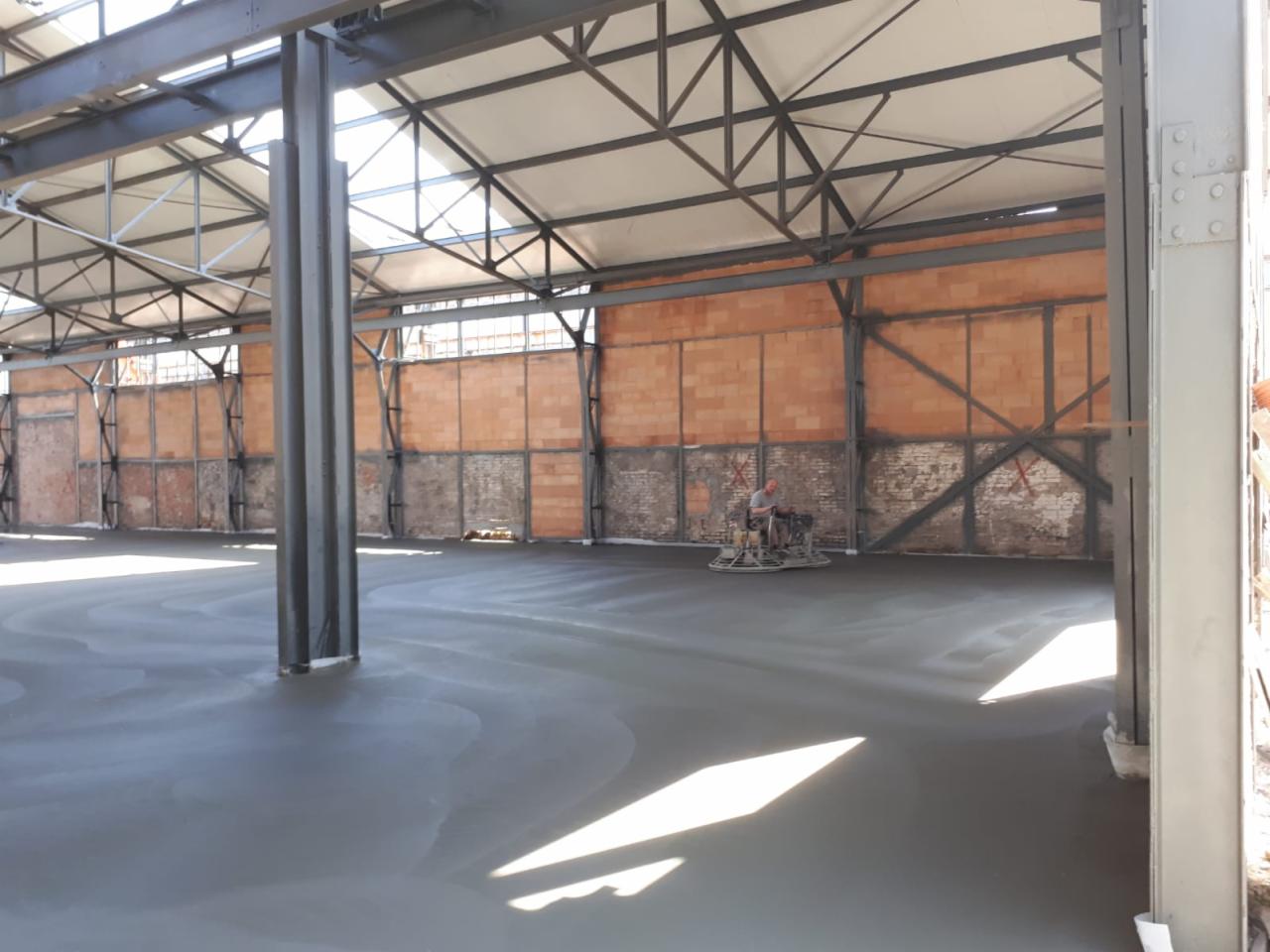 Wnętrze hali z betonową posadzką w trakcie wygładzania mechaniczną zacieraczką, widoczne metalowe słupy konstrukcyjne i murowane ściany w tle.