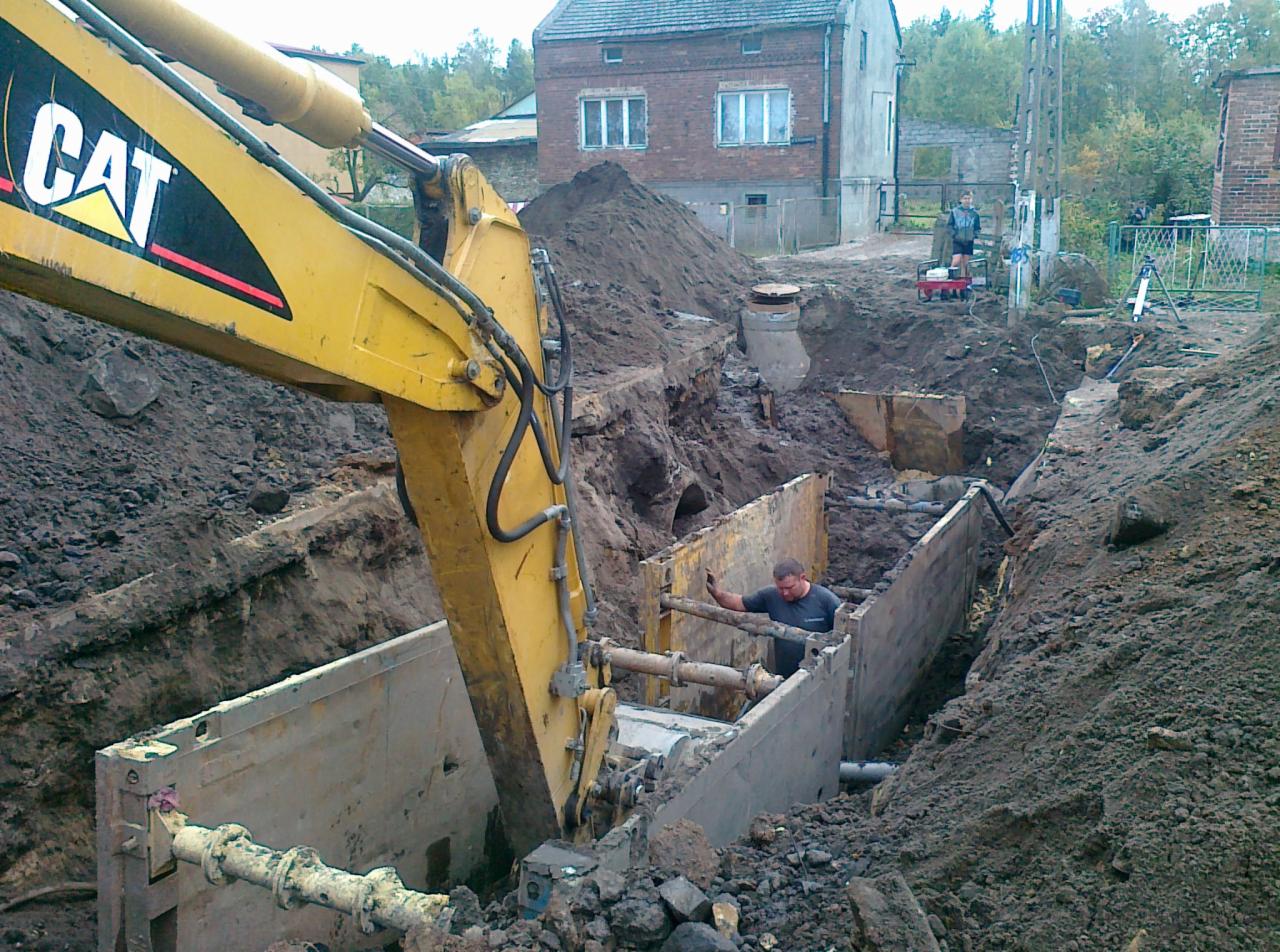 Żółta koparka CAT umieszcza betonowy element w wykopie zabezpieczonym szalunkiem, w tle budynki i hałda ziemi.