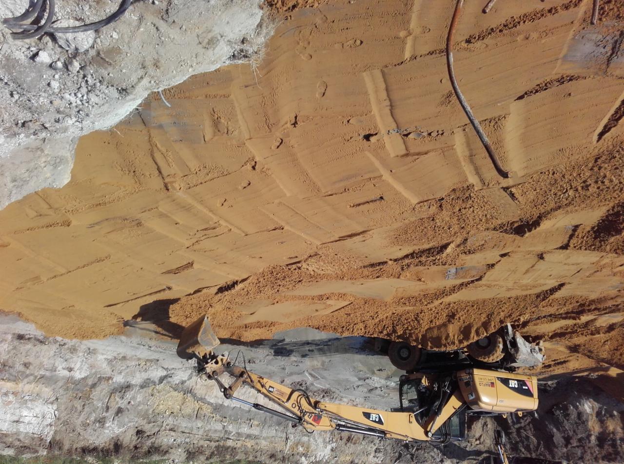 Żółta koparka CAT wybiera ziemię z wykopu o wyraźnych śladach po pracy ciężkiego sprzętu, widoczna warstwa piasku i gruzu.