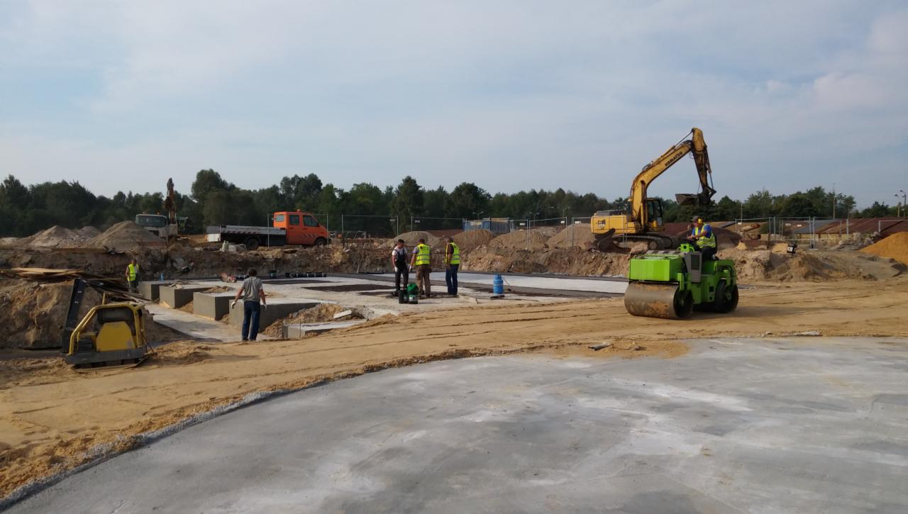 Rozległy plac budowy z wyeksponowanymi fundamentami, pracującym walcem drogowym, koparką i grupą robotników w odblaskowych kamizelkach, teren przygotowany pod utwardzenie.