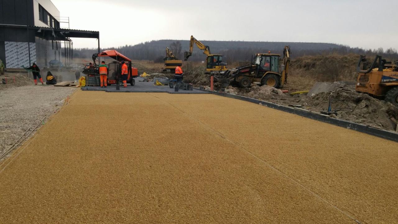 Maszyna do układania nawierzchni z kruszywa stoi na placu budowy, w tle pracują koparki i widać budynek w stanie surowym zamkniętym. Ułożona już część nawierzchni ma jasnobrązowy kolor i drobną...