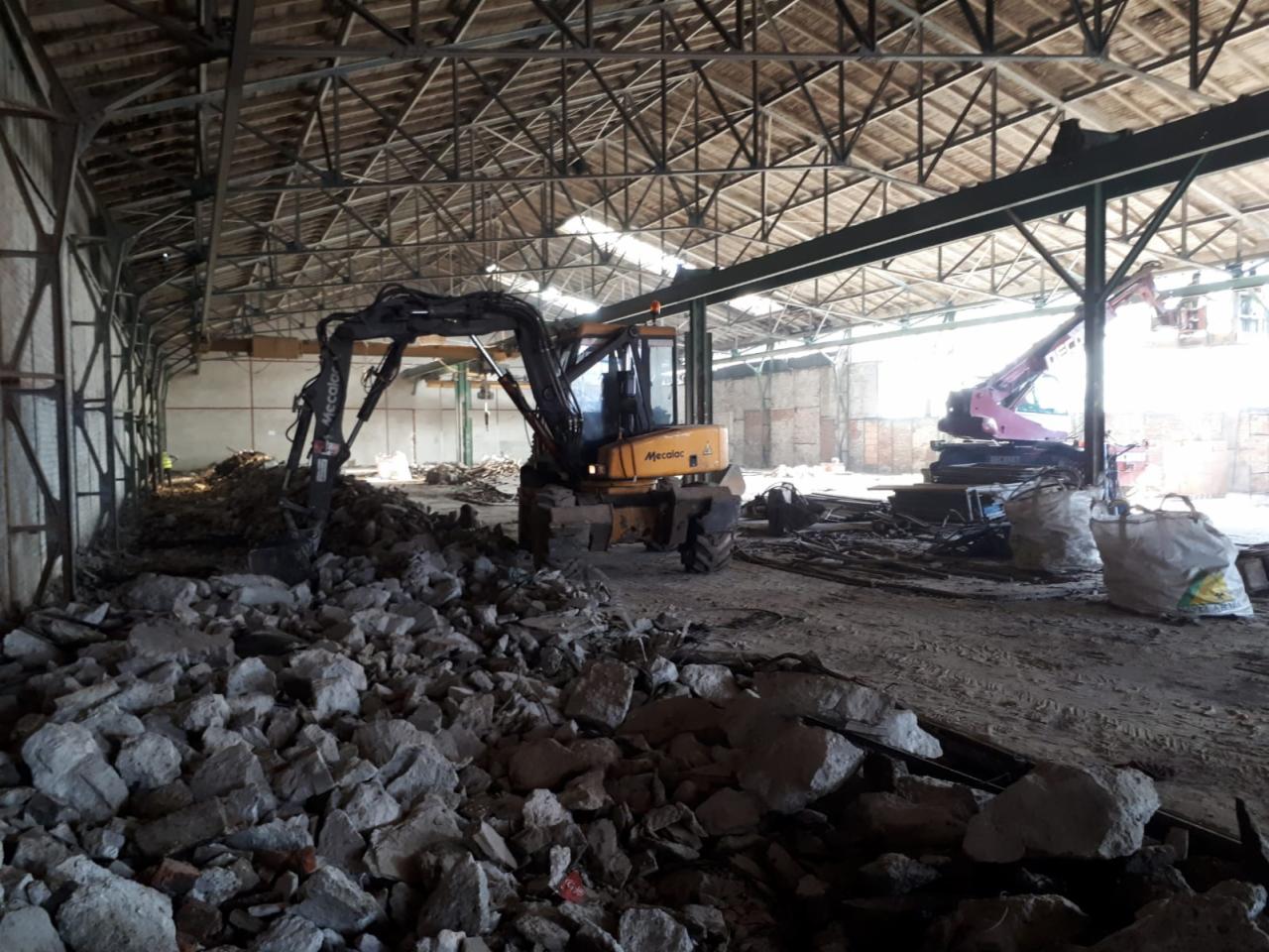 Wnętrze hali w trakcie rozbiórki z widoczną maszyną Mecalac, gruzem i elementami konstrukcji. W tle widoczny żuraw i worki.