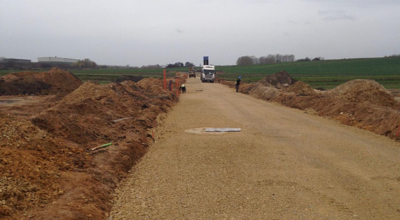 Niedokończona droga gruntowa z podsypką żwirową, widoczny ciężki sprzęt budowlany w oddali i dwóch pracowników w kaskach, po bokach hałdy ziemi.