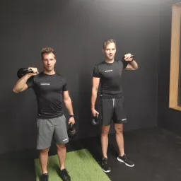 Dwaj mężczyźni w sportowych strojach ćwiczą z kettlebells w siłowni na tle ciemnej ściany i zielonego dywanika.