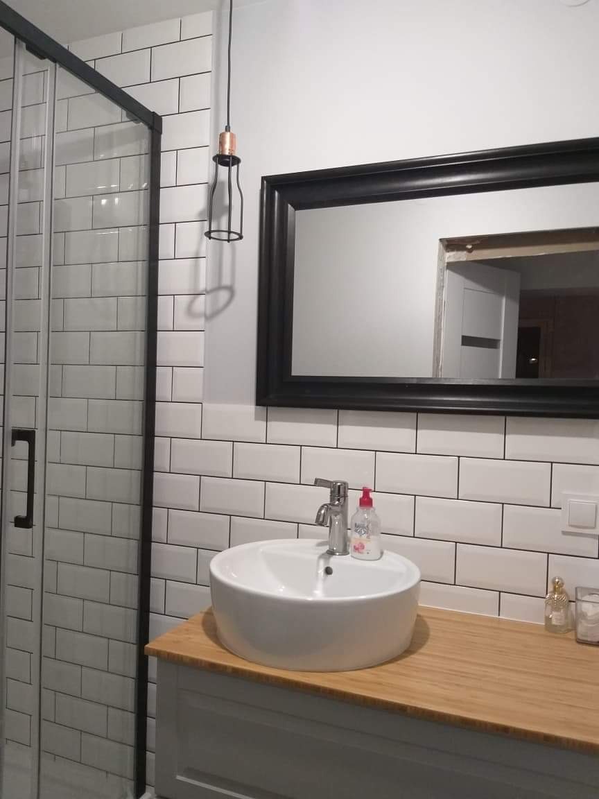 Wykończona łazienka z kabiną prysznicową z czarnymi ramami, białą umywalką nablatową na drewnianym blacie, białymi płytkami typu metro i czarnym lustrem.