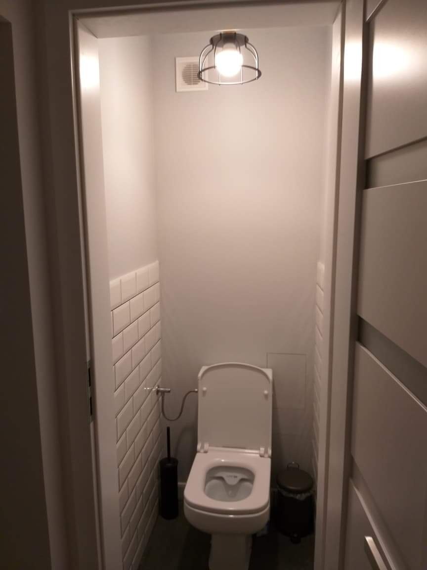 Wąska toaleta po remoncie, z białymi płytkami ułożonymi cegiełkowo do połowy ściany, wiszącą lampą w stylu industrialnym i szarą podłogą.