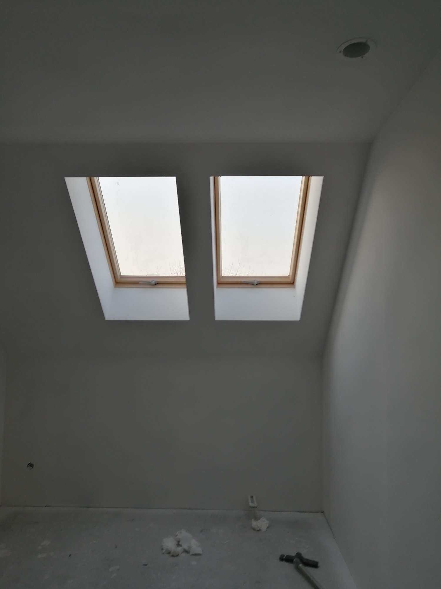 Wnętrze pokoju na poddaszu z dwoma oknami dachowymi z drewnianymi ramami i białymi ścianami, widoczne ślady prac wykończeniowych na podłodze.