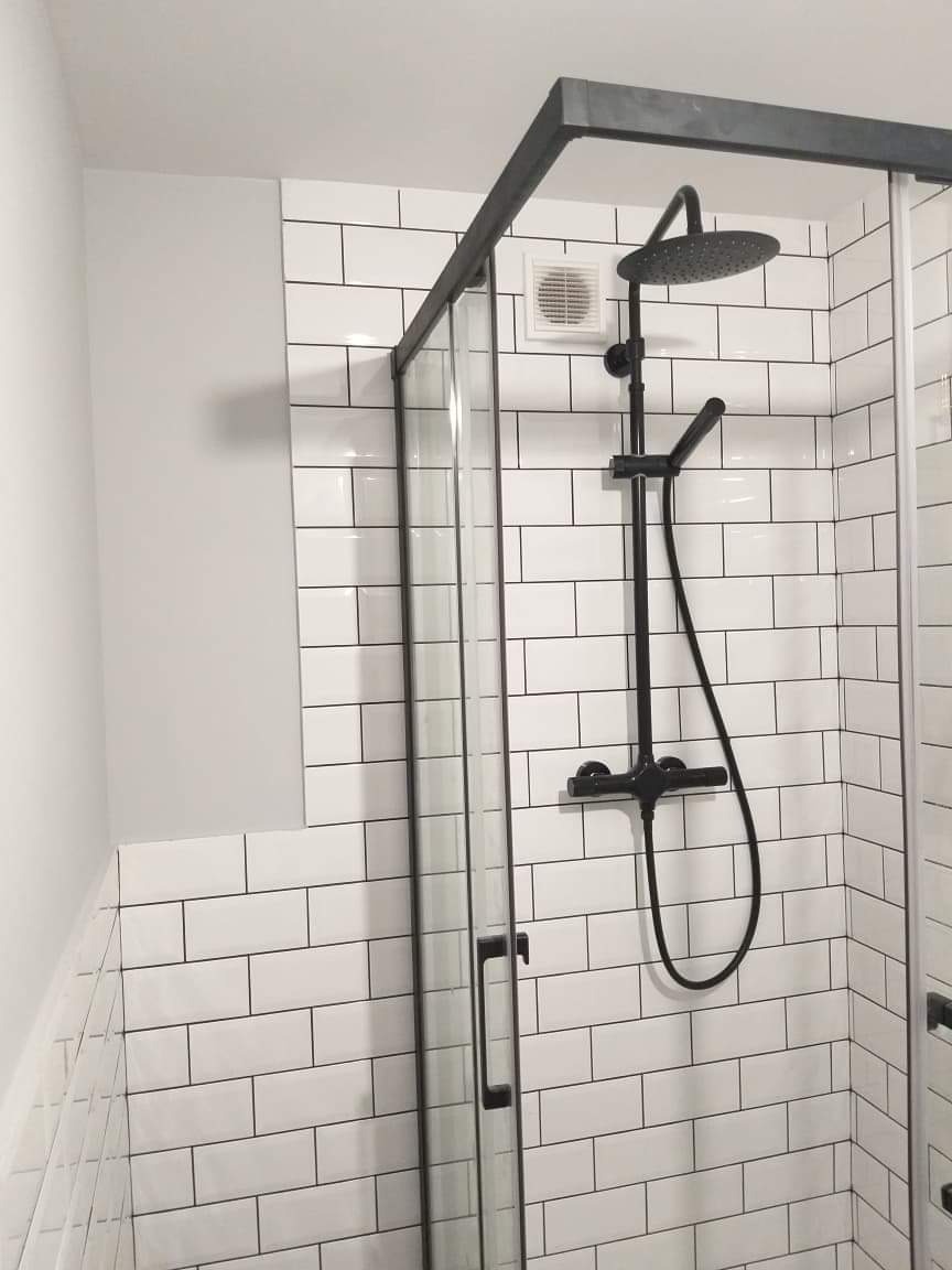 Kabina prysznicowa z czarną armaturą na tle białych, prostokątnych płytek, widoczny wentylator w ścianie.