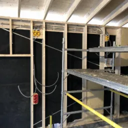 Wnętrze w trakcie budowy: drewniany szkielet ściany z czarną membraną, instalacja elektryczna w peszlach, fragment rusztowania i wełna mineralna między słupkami.