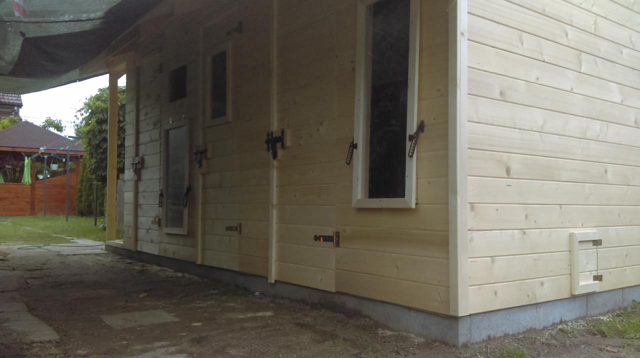 Zewnętrzny widok drewnianej konstrukcji z jasnej tarcicy, z oknami i okuciami, zadaszona plandeką, na betonowej podmurówce.