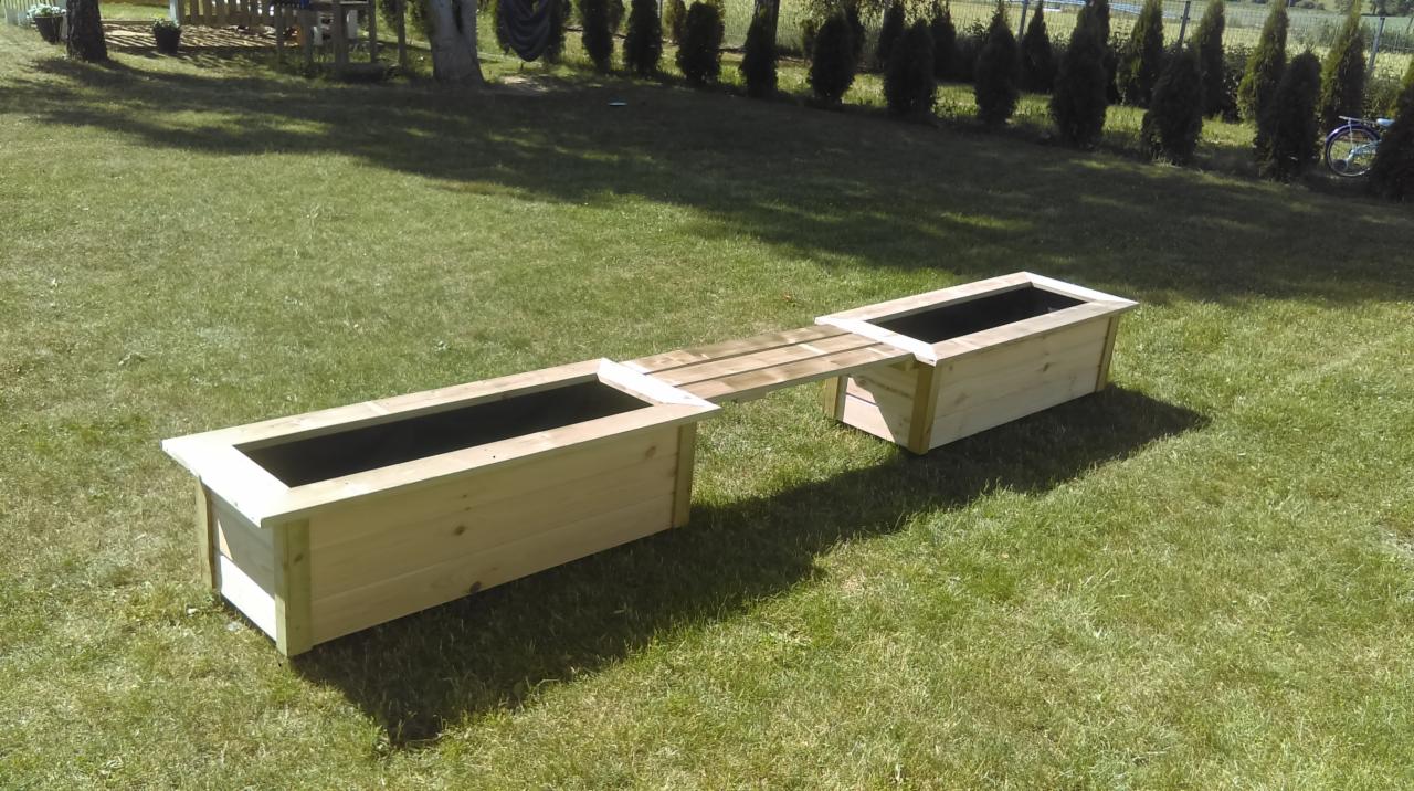 Dwa drewniane koryta na rośliny połączone drewnianym mostkiem, stojące na trawniku w ogrodzie.