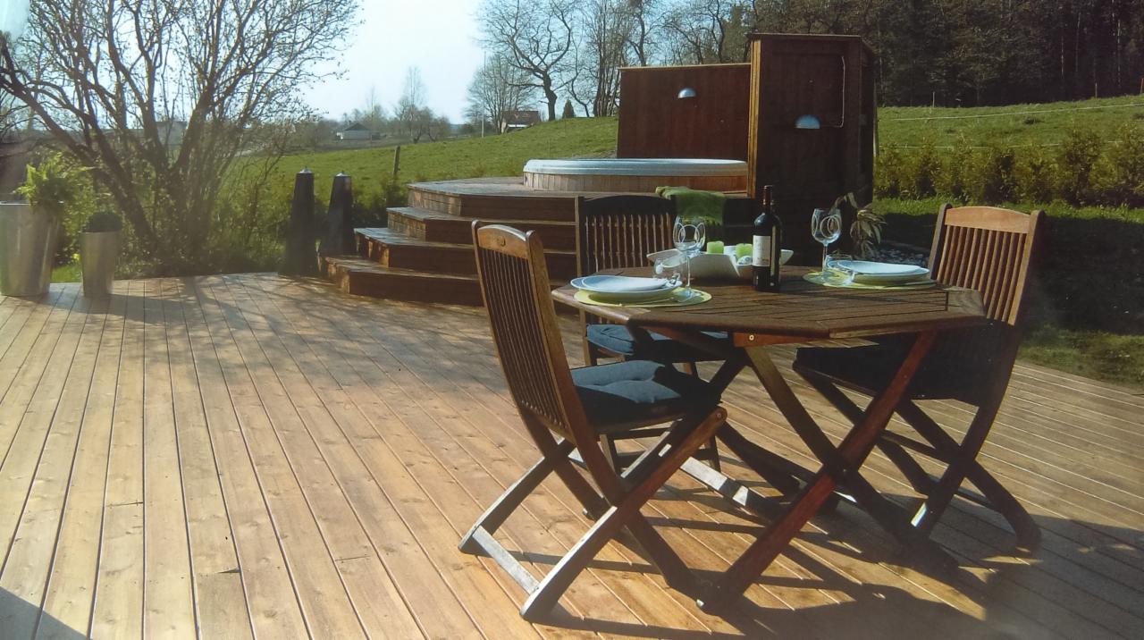 Drewniany taras z jacuzzi na podwyższeniu, stół z nakryciem i krzesłami, butelka wina, widok na zielony trawnik i drzewa w tle.