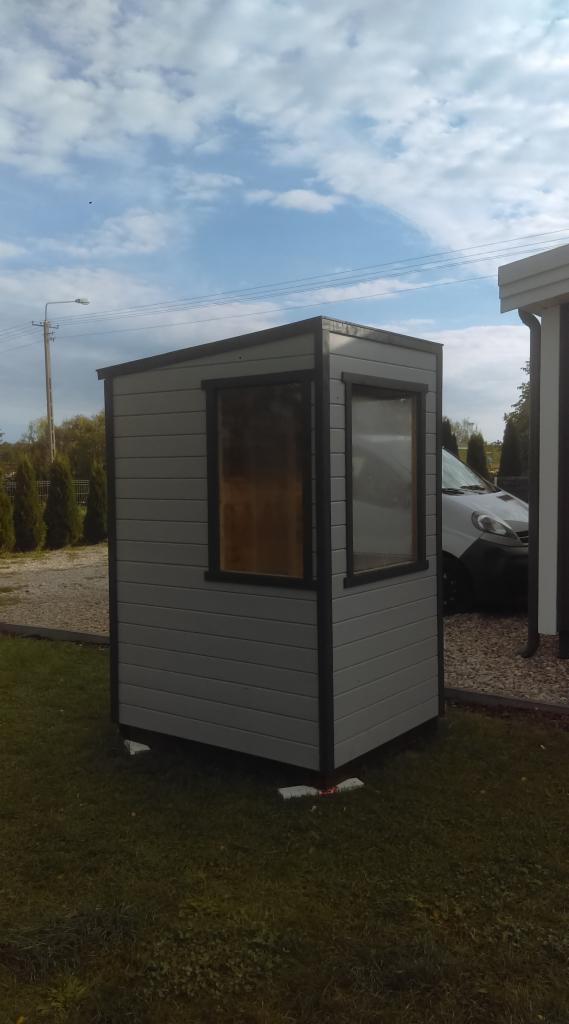 Gotowa budka z szarej deski elewacyjnej z dwoma oknami, czarnymi obramowaniami i dachem, umieszczona na trawniku, z widocznym samochodem w tle.