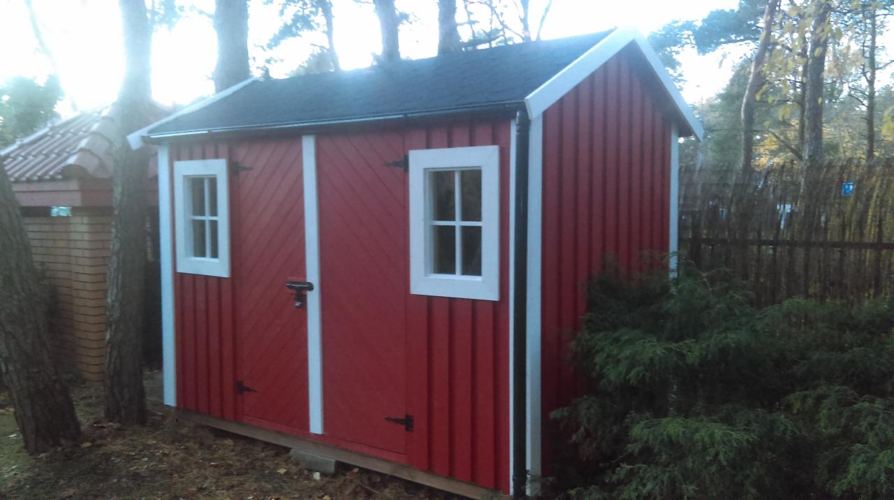 Czerwona, drewniana szopa narzędziowa z białymi obramowaniami okien i drzwi, czarnym dachem i prostym zamkiem, otoczona drzewami i krzewami.